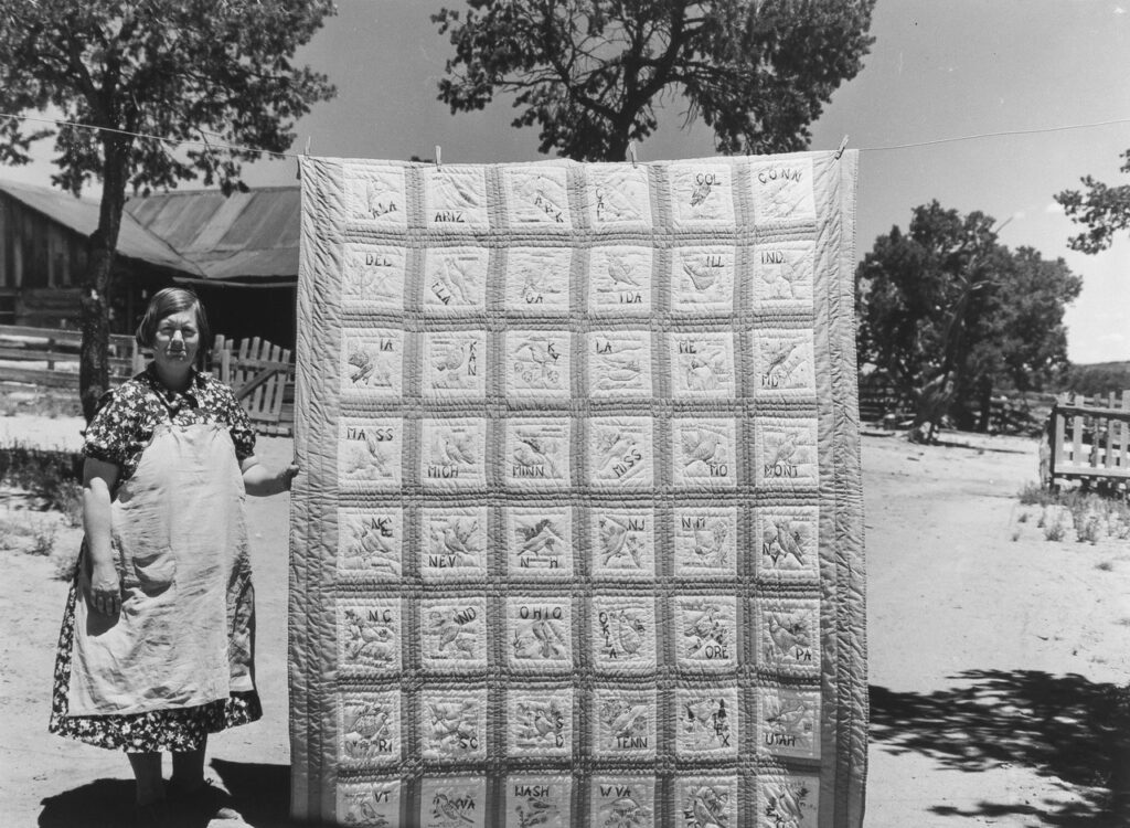 Russell Lee (1903 - 1986). A community settles by about 200 migrant Texas and Oklahoma farmers who filed homestead claims. Mrs. Bill Stagg with state quilt which she made. Mrs. Staggs helps her husband in the fields with plowing, planting, weeding corn and harvesting beans. She quilts while she rests during the noon hour. Pie Town, New Mexico. June 1940.
