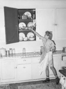 Russell Lee (1903 - 1986). Daughter of mormon farmer putting dishes in kitchen cabinet. Box Elder County, Utah. August 1940.