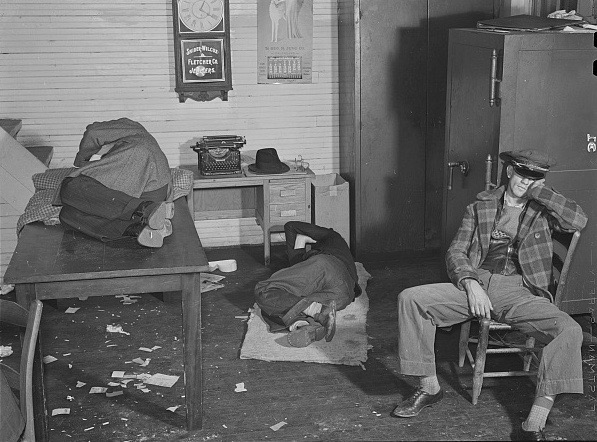 Marion Post Wolcott (1910 - 1990). Farmers must often wait over night before their tobacco is sold at auction. They sleep anywhere, one is lying on a desk, one on the floor, and one is sitting in a chair sleeping in this warehouse office. Durham, North Carolina. November 1939